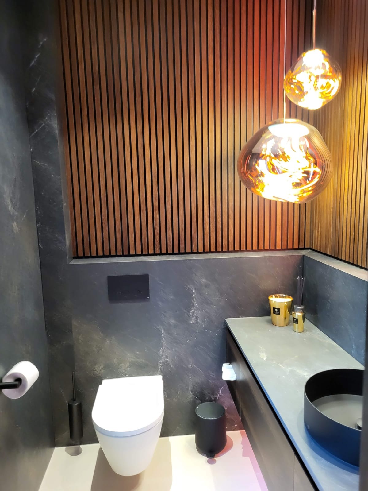 Nowoczesna łazienka z drewnianą ścianą, marmurowymi płytami, podwieszaną toaletą i dekoracyjnymi lampami wiszącymi w bursztynowym kolorze.