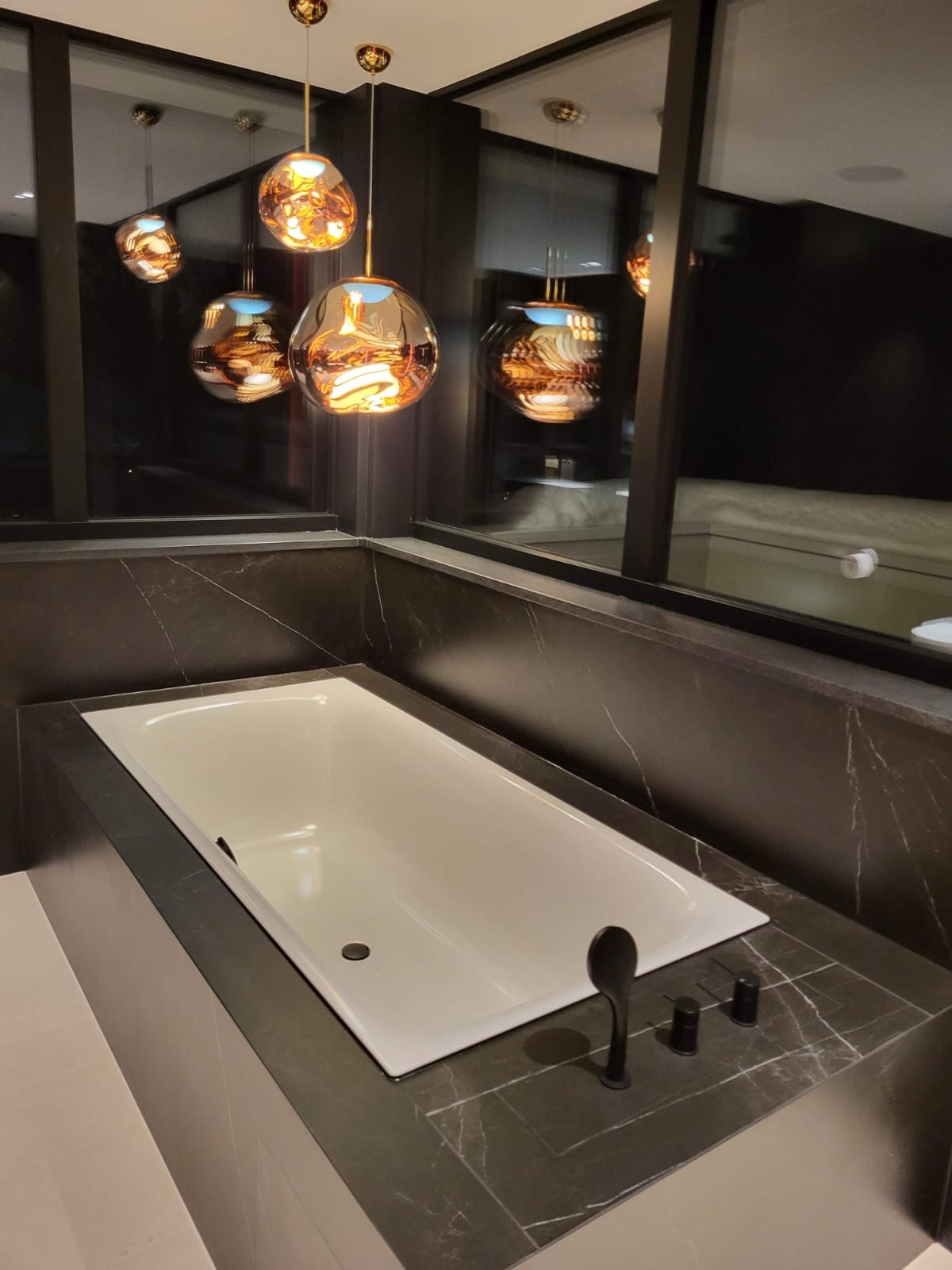 Biała wanna obudowana czarnym marmurem z widoczną czarną armaturą, oświetlona dekoracyjnymi, złotymi lampami wiszącymi, odbijającymi się w szybach okien.