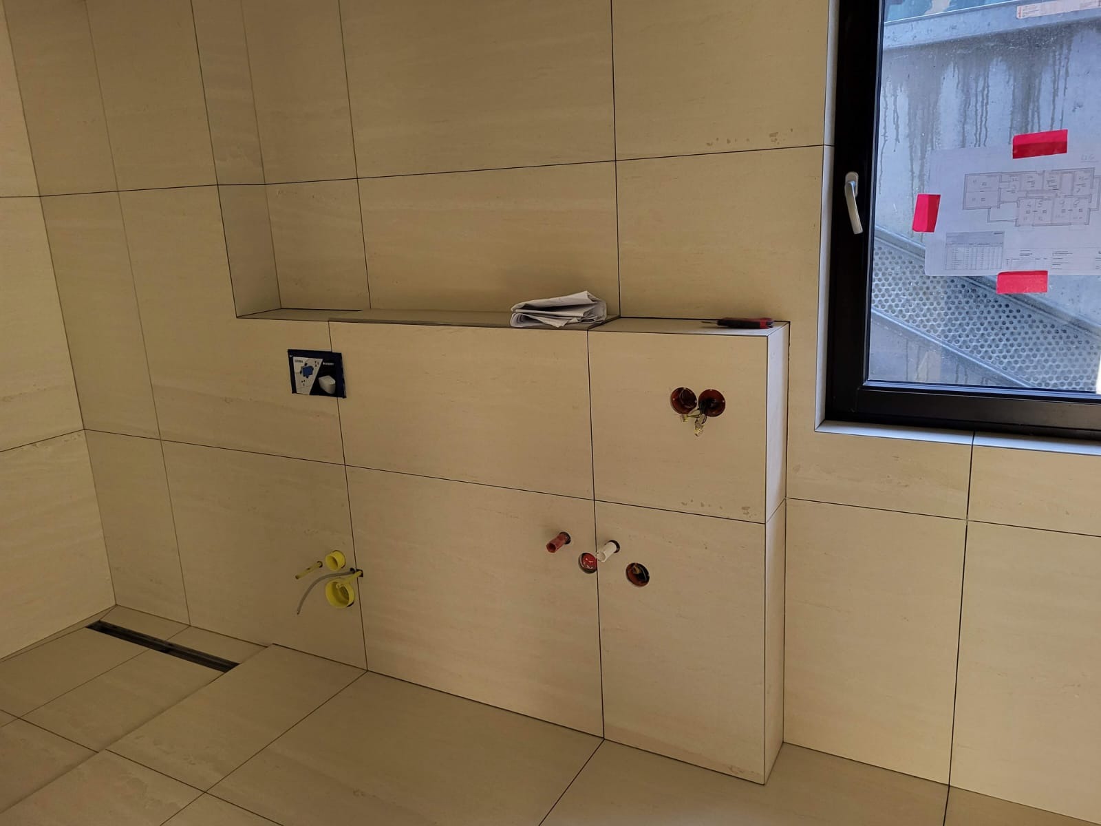 Wnętrze łazienki w trakcie wykańczania: beżowe płytki imitujące kamień, widoczne instalacje hydrauliczne w ścianie, okno z widokiem na schody zewnętrzne, plan architektoniczny przyklejony do szyby.