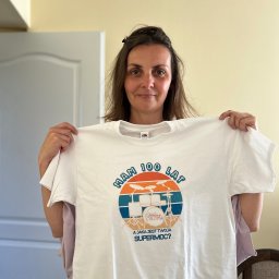 About Image - Uśmiechnięta kobieta trzyma białą koszulkę z nadrukiem: 'Mam 100 lat', grafika z perkusją i napis 'A jaka jest Twoja supermoc?' na beżowym tle.