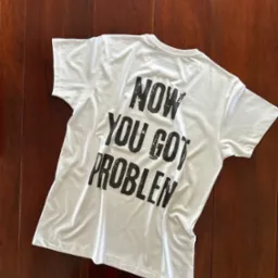 Biała koszulka z czarnym nadrukiem 'NOW YOU GOT PROBLEM' leży na ciemnobrązowym, drewnianym podłożu. Widok z góry, oświetlenie naturalne.