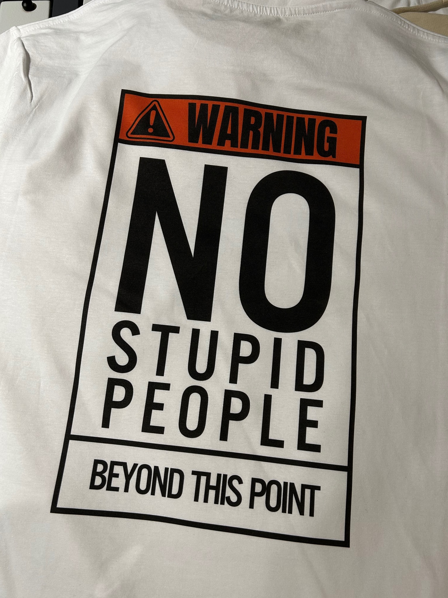 Biała koszulka z czarnym nadrukiem: 'WARNING NO STUPID PEOPLE BEYOND THIS POINT'. Nadruk imituje znak ostrzegawczy.