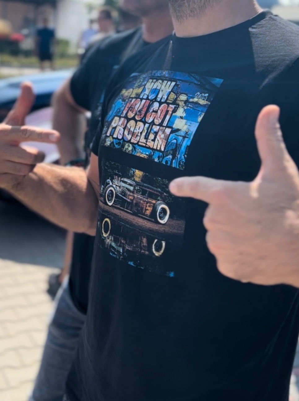 Mężczyzna w czarnej koszulce z nadrukiem oldschoolowego samochodu i napisem 'NOW YOU GOT PROBLEM'. Fragmenty sylwetek innych osób w tle, ujęcie z bliska.
