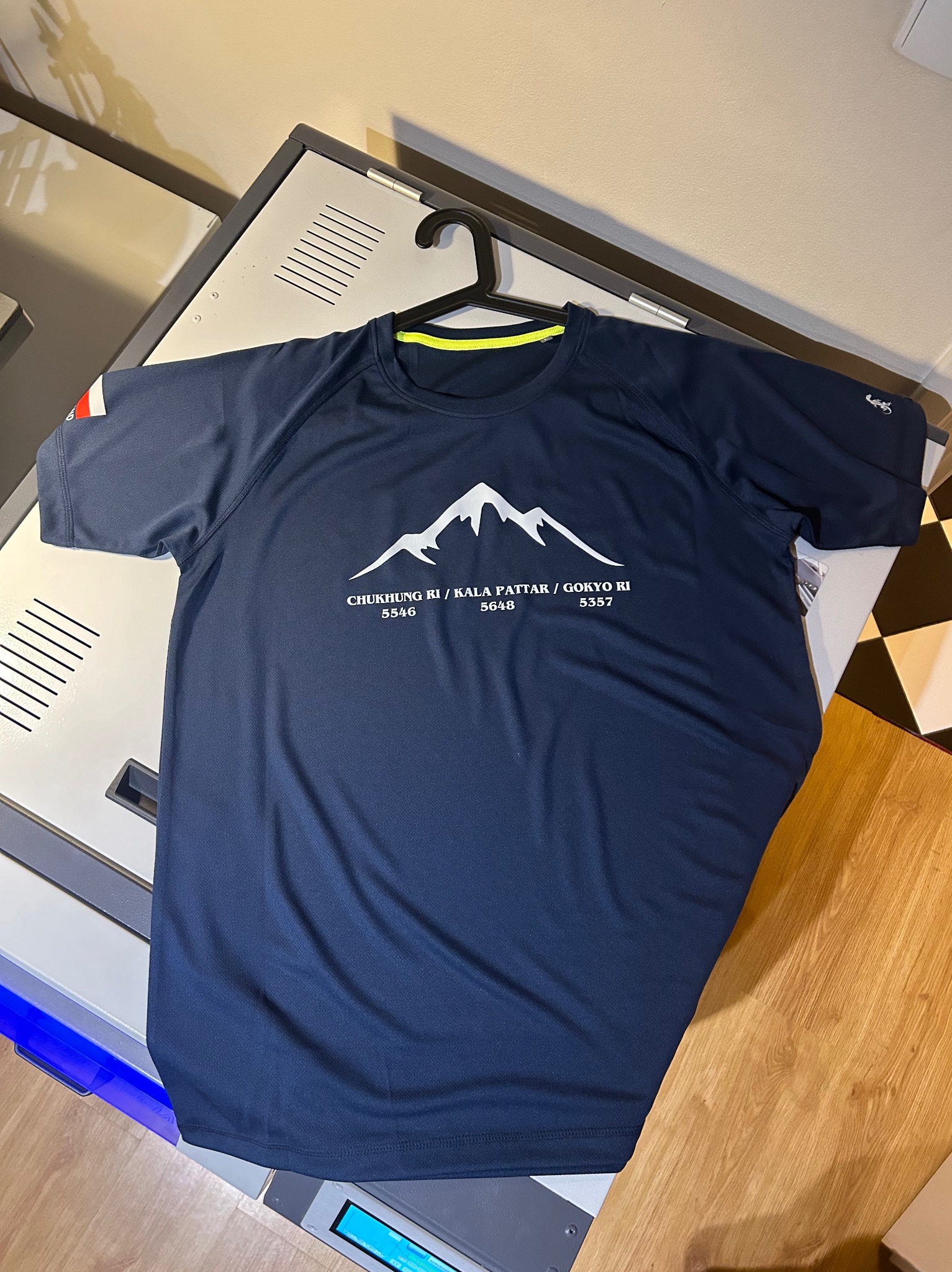 Granatowa koszulka sportowa z białym nadrukiem górskim, zawieszona na wieszaku, prezentowana na tle blatu z elementami metalowymi i drewnianą podłogą.