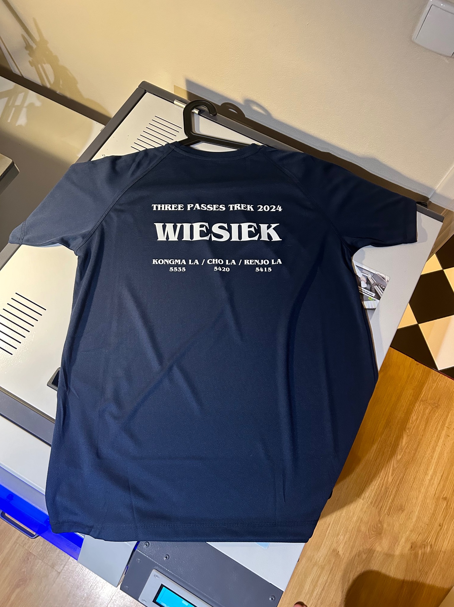 Granatowa koszulka z białym nadrukiem 'THREE PASSES TREK 2024 WIESIEK KONGMA LA / CHO LA / RENJO LA' wisi na wieszaku, leżąc na urządzeniu drukarskim.