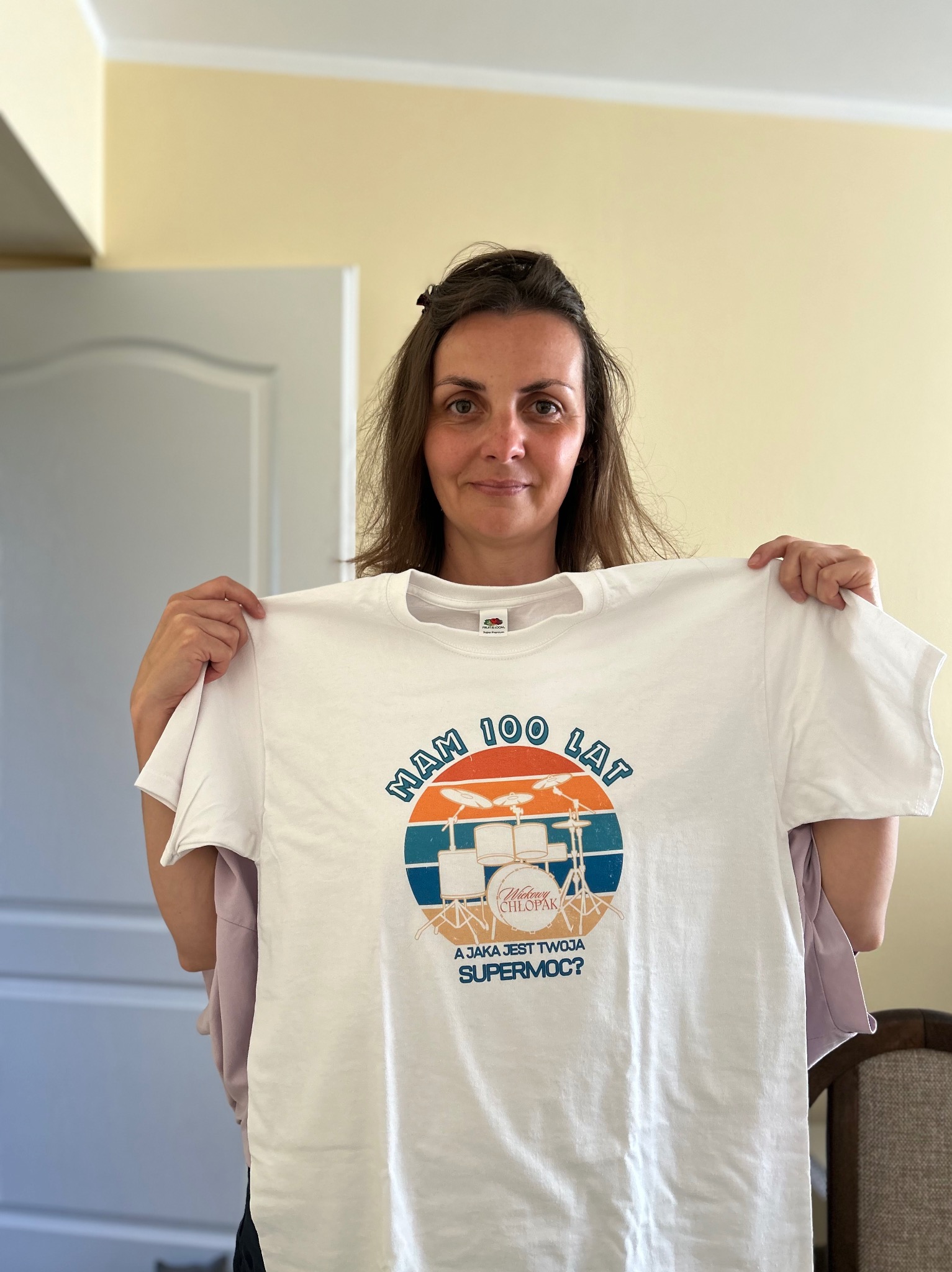 Uśmiechnięta kobieta trzyma białą koszulkę z nadrukiem: 'Mam 100 lat', grafika z perkusją i napis 'A jaka jest Twoja supermoc?' na beżowym tle.