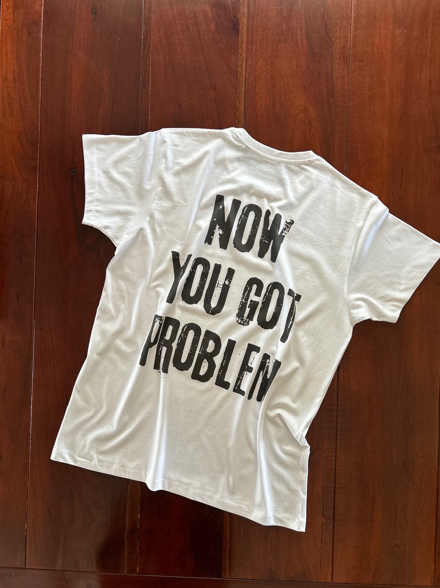 Biała koszulka z czarnym nadrukiem 'NOW YOU GOT PROBLEM' leży na ciemnobrązowym, drewnianym podłożu. Widok z góry, oświetlenie naturalne.
