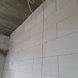 P.H.U KINA - Biała ściana z betonu komórkowego w trakcie budowy, z poprowadzonym kablem elektrycznym zabezpieczonym uchwytami. Kabel zwisa luźno z sufitu. Widoczny fragment surowego betonowego stropu.