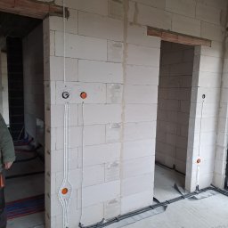 P.H.U KINA - Surowy stan: Białe ściany z betonu komórkowego w trakcie budowy, widoczne prowadzone przewody elektryczne w peszlach i rurkach, puszki instalacyjne w kolorze pomarańczowym, brak wykończenia podłóg...