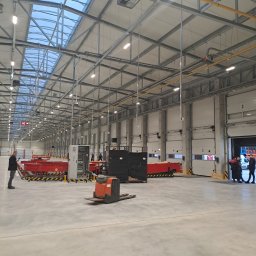 P.H.U KINA - Wnętrze hali magazynowej z widocznymi instalacjami pod sufitem, oświetleniem LED, skrzynką elektryczną i pracownikami przy bramie wjazdowej.