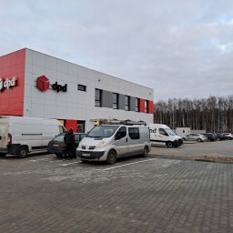 P.H.U KINA - Srebrny van z drabinką na dachu zaparkowany na parkingu przed budynkiem DPD, obok stoją inne samochody, a w tle widoczny las i słupy wysokiego napięcia.