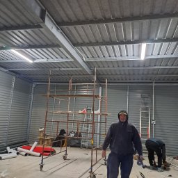 P.H.U KINA - Pracownik z papierosem w ustach, w ubraniu roboczym, w hali z blachy falistej, na pierwszym planie rusztowanie, na drugim planie drabina i drugi pracownik przy podłodze, widoczne rury i kable.