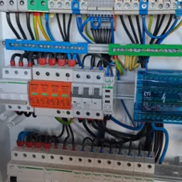 Wnętrze skrzynki rozdzielczej z zainstalowanymi bezpiecznikami, ogranicznikami przepięć i przewodami o różnych kolorach, widoczne elementy montażowe i oznaczenia producentów.