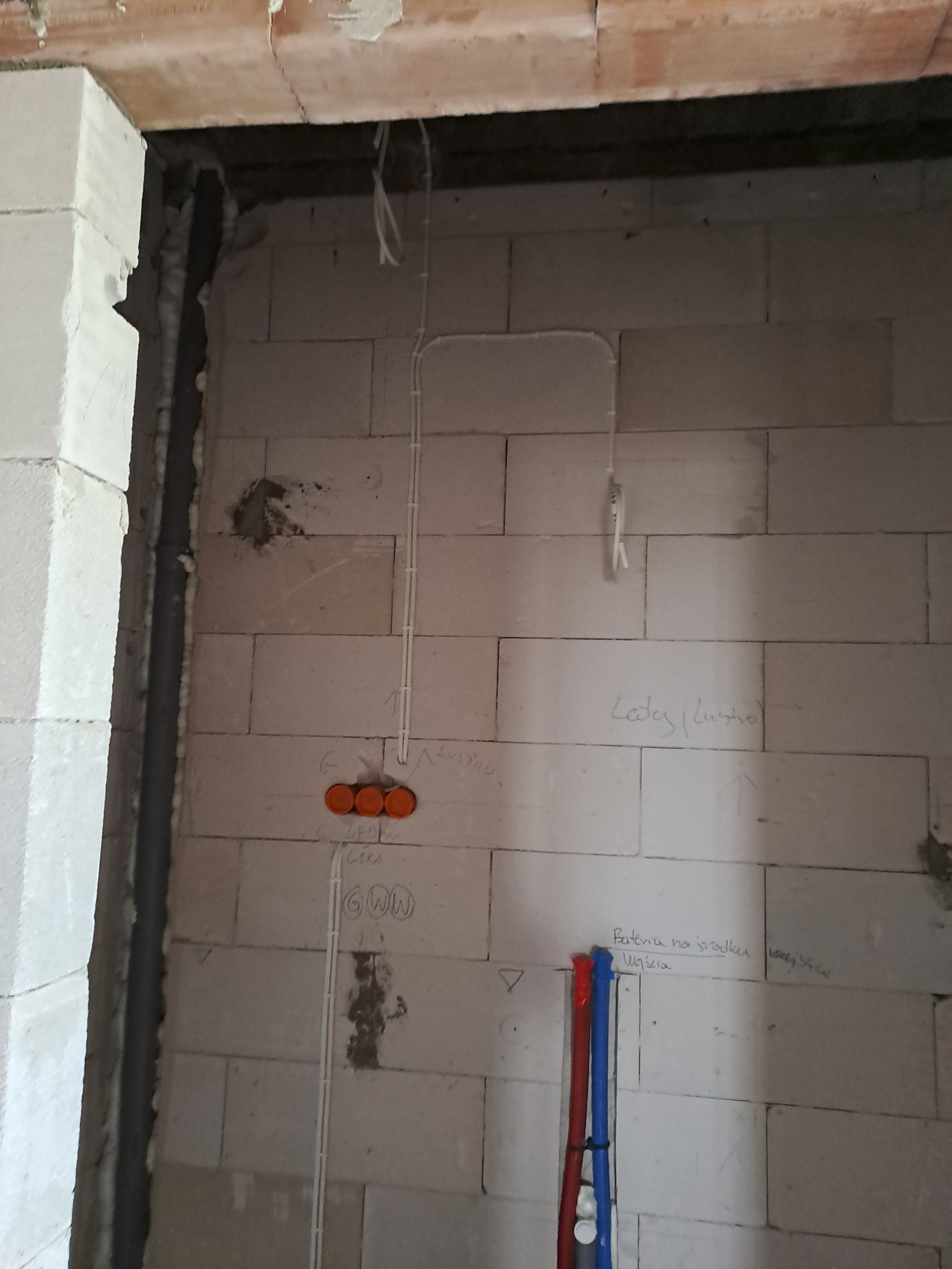 Surowa ściana z betonu komórkowego z widocznymi przewodami elektrycznymi w peszlach, puszkami instalacyjnymi z pomarańczowymi dekielkami, instalacją wodną w kolorach czerwonym i niebieskim...