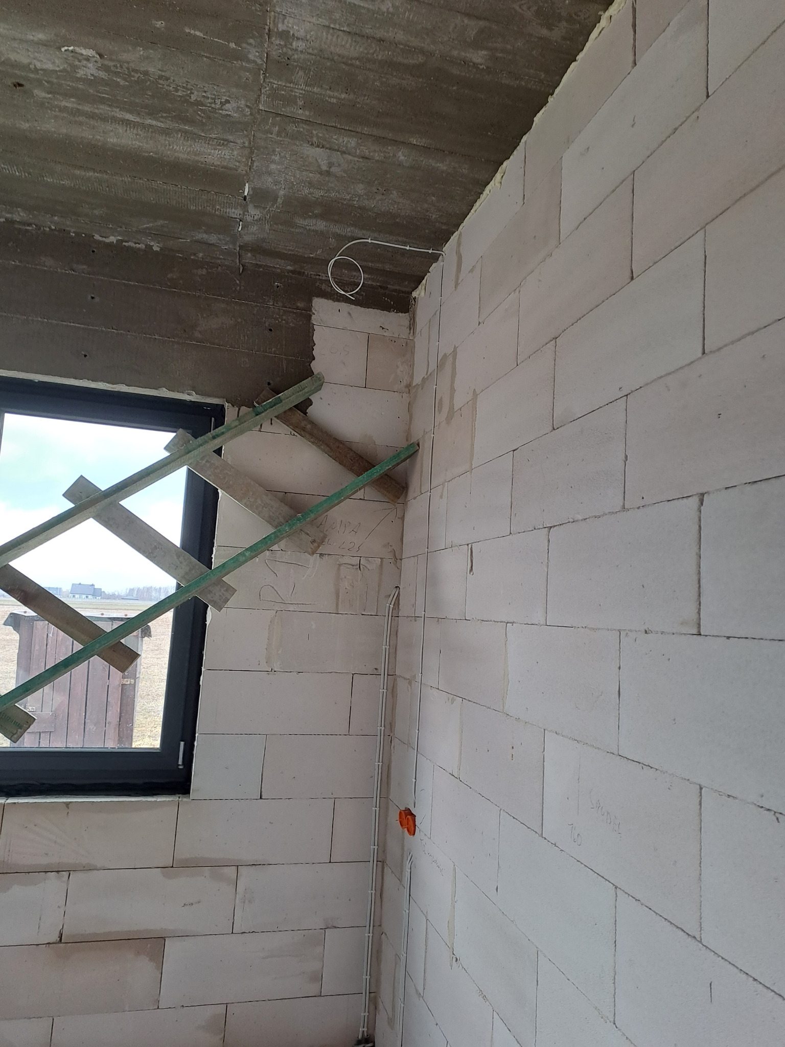 Surowy stan wnętrza budynku w trakcie budowy: ściany z białych bloczków, okno z drewnianą konstrukcją zabezpieczającą, widoczne przewody elektryczne przy ścianie i pod sufitem.