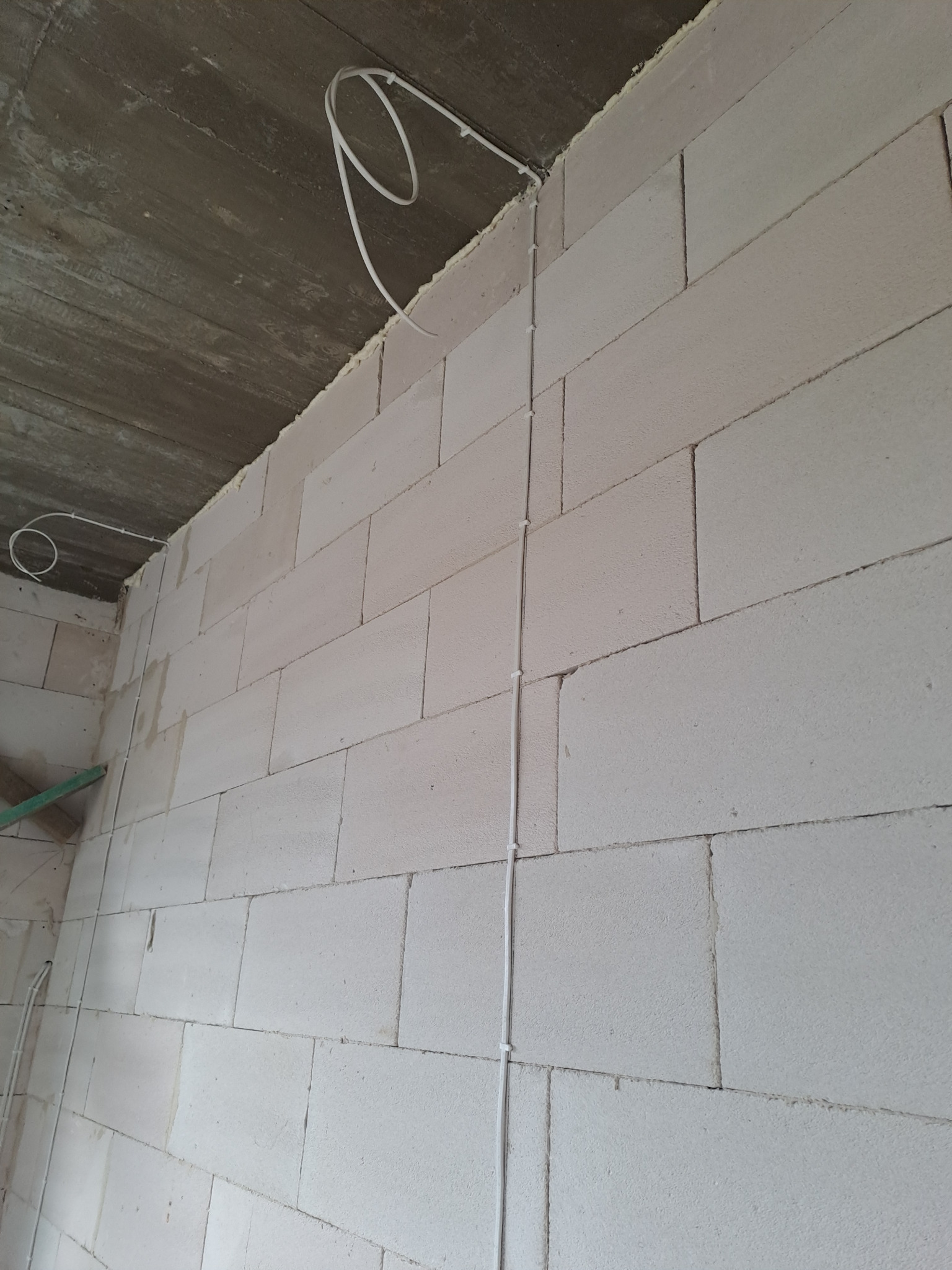 Biała ściana z betonu komórkowego w trakcie budowy, z poprowadzonym kablem elektrycznym zabezpieczonym uchwytami. Kabel zwisa luźno z sufitu. Widoczny fragment surowego betonowego stropu.