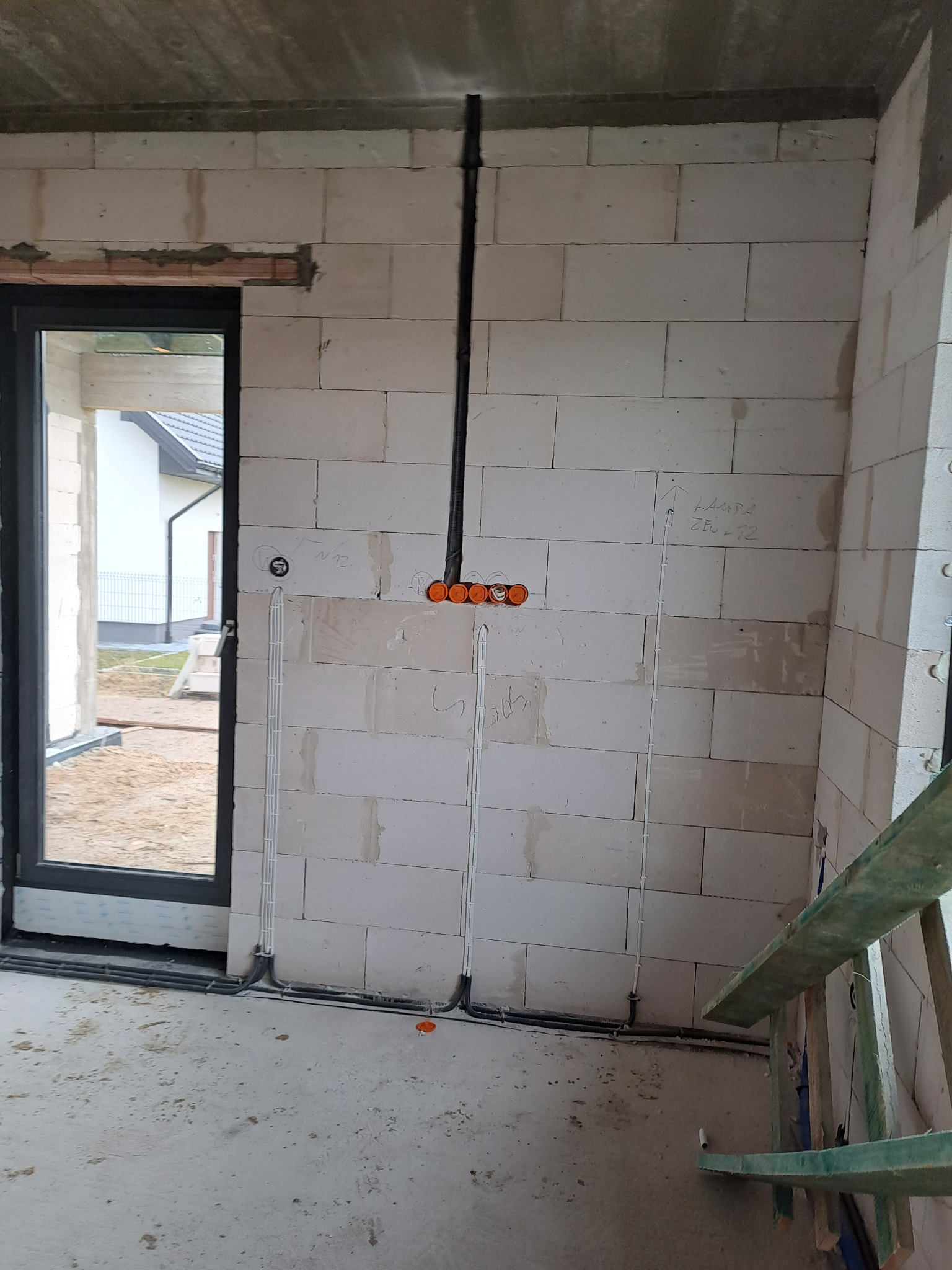 Surowy stan wnętrza z widocznymi przewodami elektrycznymi prowadzonymi po ścianie z białego bloczka, puszki instalacyjne pomarańczowe, oznaczenia na ścianie, w tle widok przez okno na budynek...