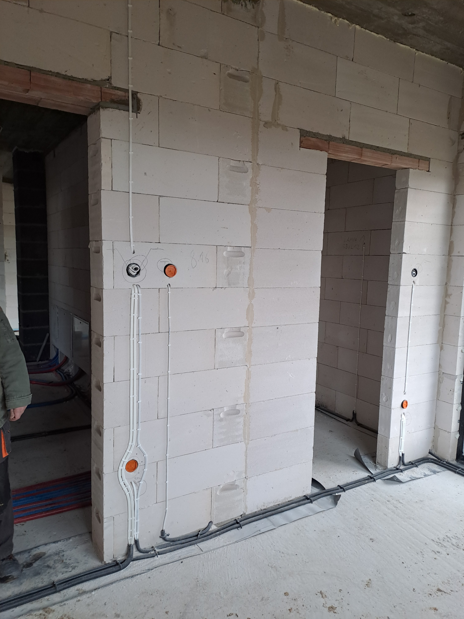 Surowy stan: Białe ściany z betonu komórkowego w trakcie budowy, widoczne prowadzone przewody elektryczne w peszlach i rurkach, puszki instalacyjne w kolorze pomarańczowym, brak wykończenia podłóg...