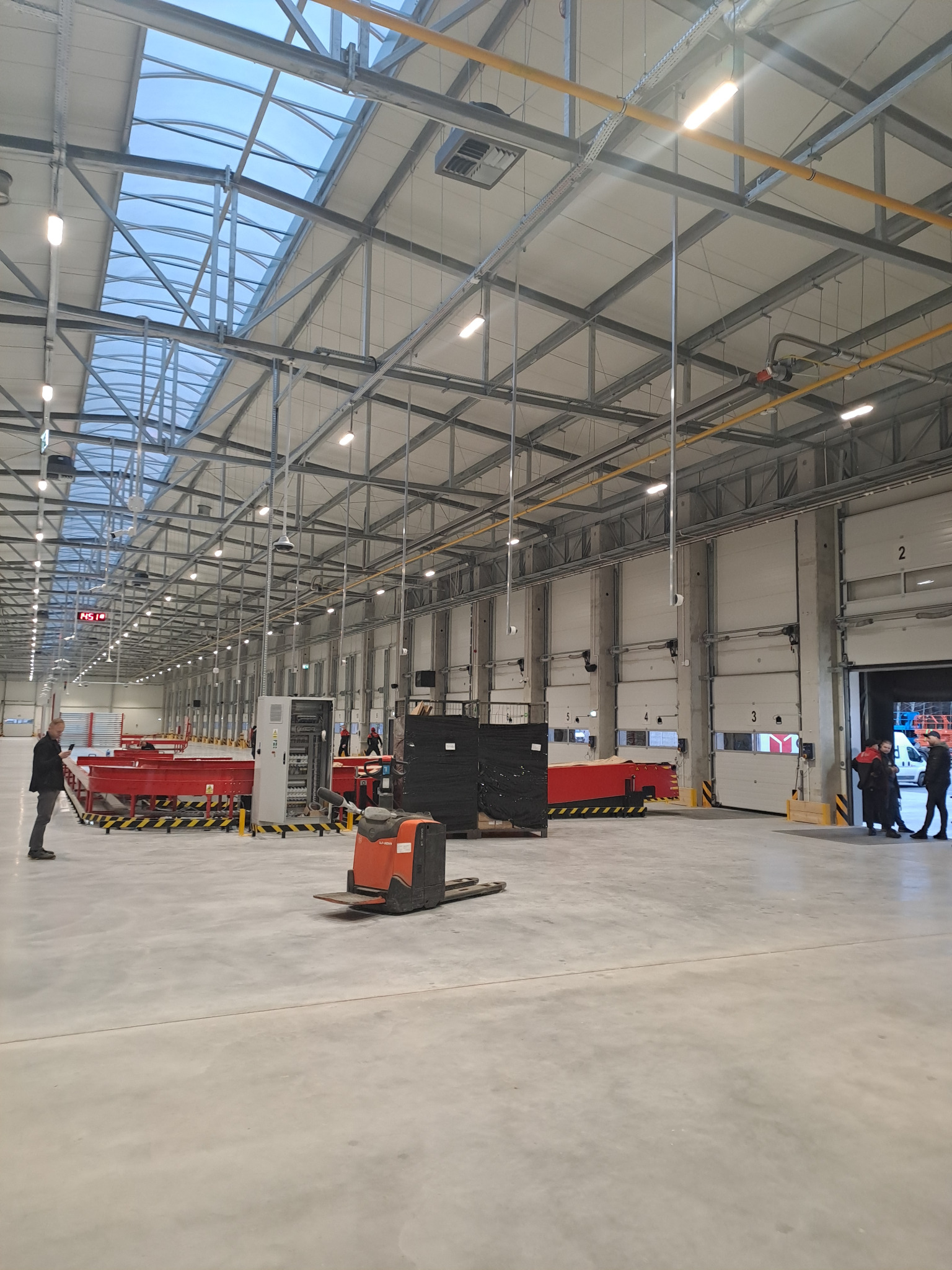 Wnętrze hali magazynowej z widocznymi instalacjami pod sufitem, oświetleniem LED, skrzynką elektryczną i pracownikami przy bramie wjazdowej.