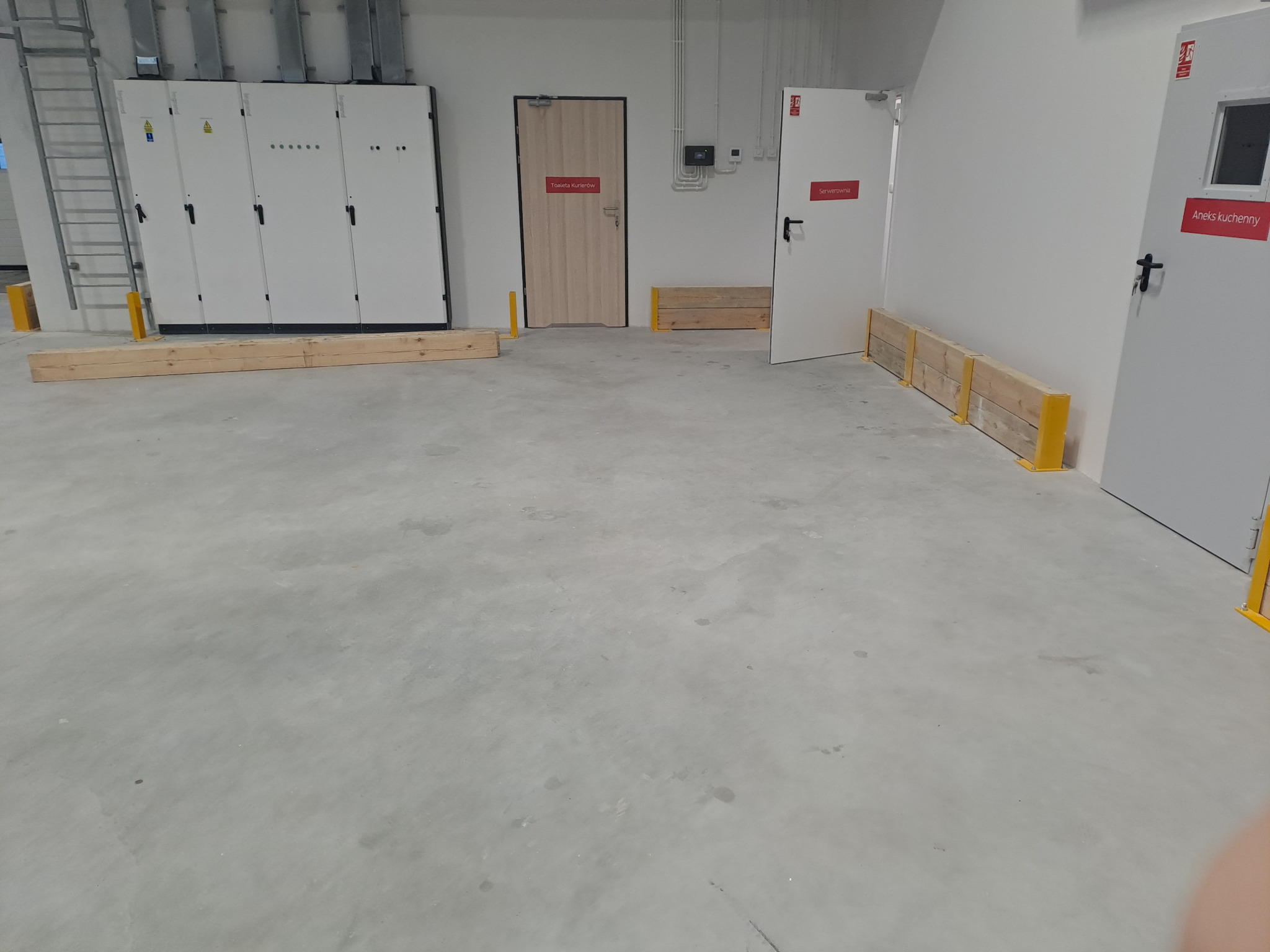 Wnętrze techniczne z rozdzielnicą elektryczną, zabezpieczone drewnianymi barierami, widoczne drzwi do serwerowni i toalety kurierów.