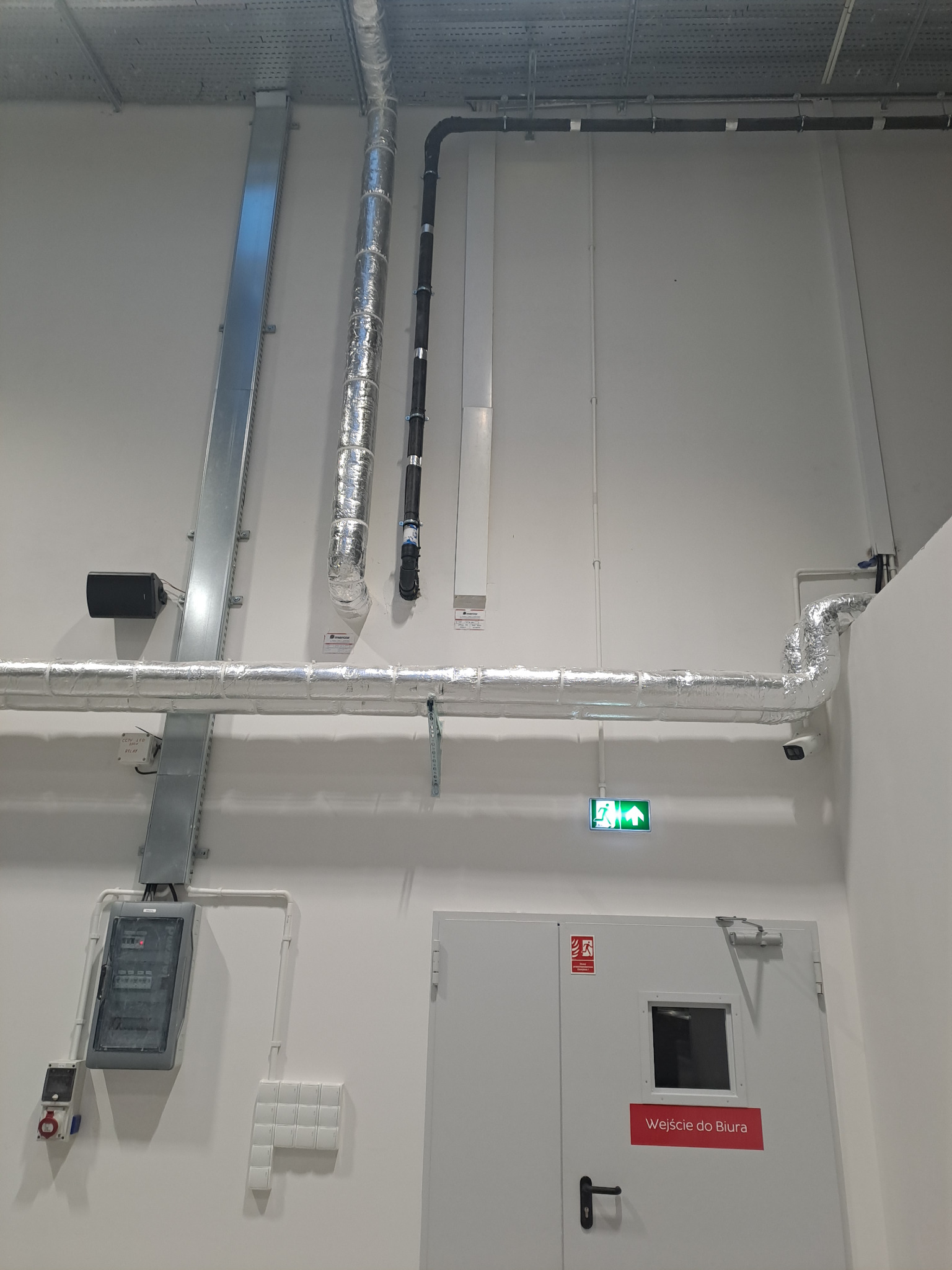 Rozbudowana instalacja elektryczna i wentylacyjna w obiekcie biurowym w Szydłowcu, widoczne koryta kablowe, rury w izolacji, tablica rozdzielcza i oświetlenie awaryjne nad drzwiami.