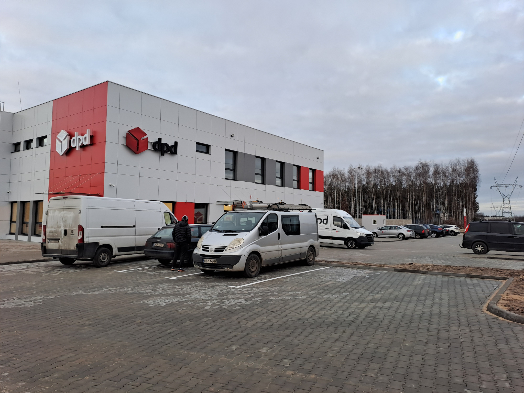 Srebrny van z drabinką na dachu zaparkowany na parkingu przed budynkiem DPD, obok stoją inne samochody, a w tle widoczny las i słupy wysokiego napięcia.