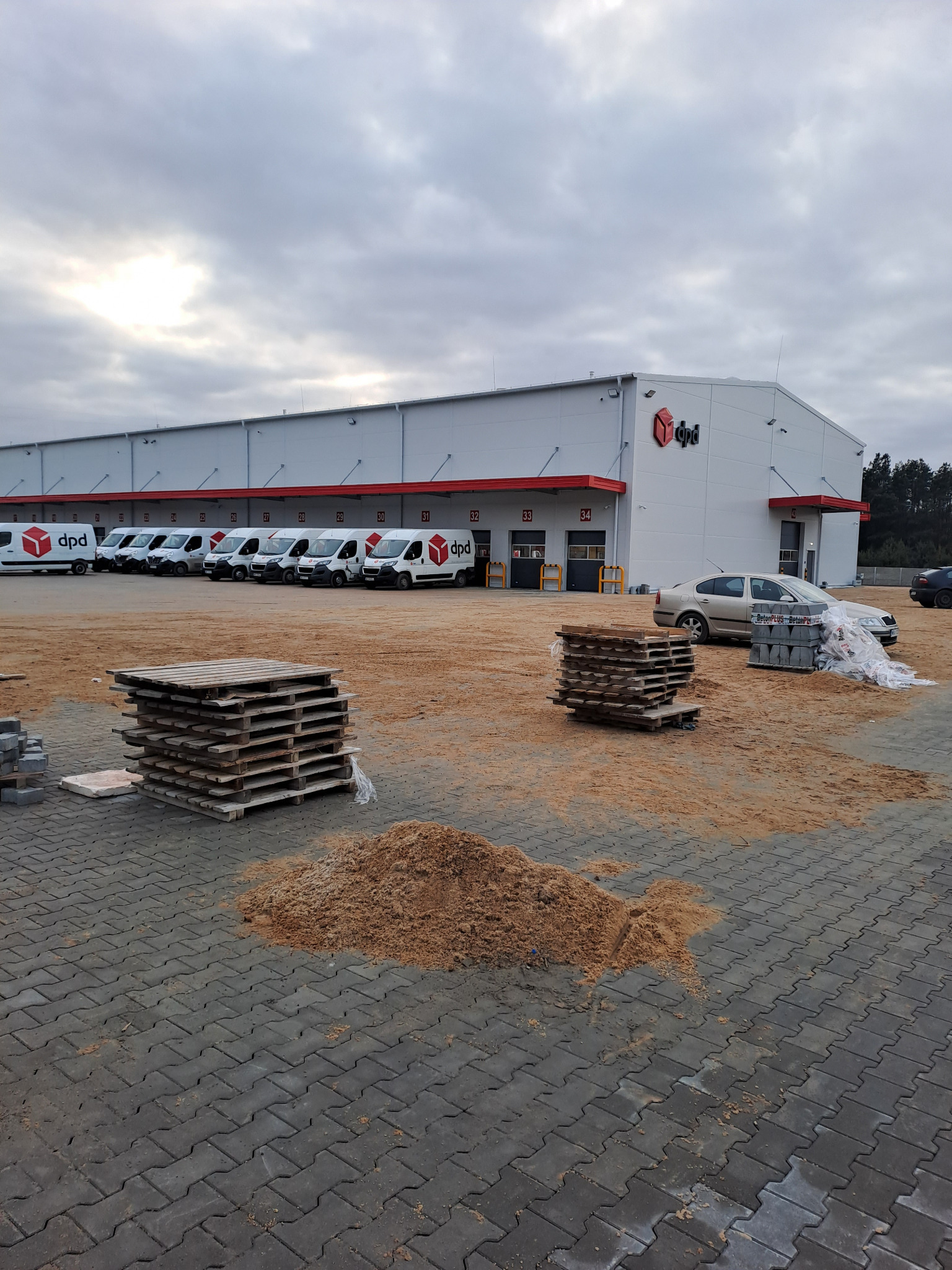 Plac budowy przed magazynem firmy kurierskiej DPD z ułożoną kostką brukową, paletami i stertą piasku. W tle zaparkowane samochody dostawcze DPD.