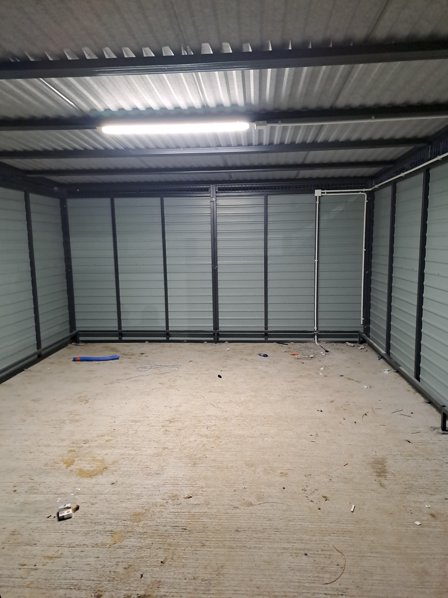 Wnętrze metalowego garażu z widoczną instalacją elektryczną: biała lampa LED pod sufitem i białe kable poprowadzone wzdłuż ściany.