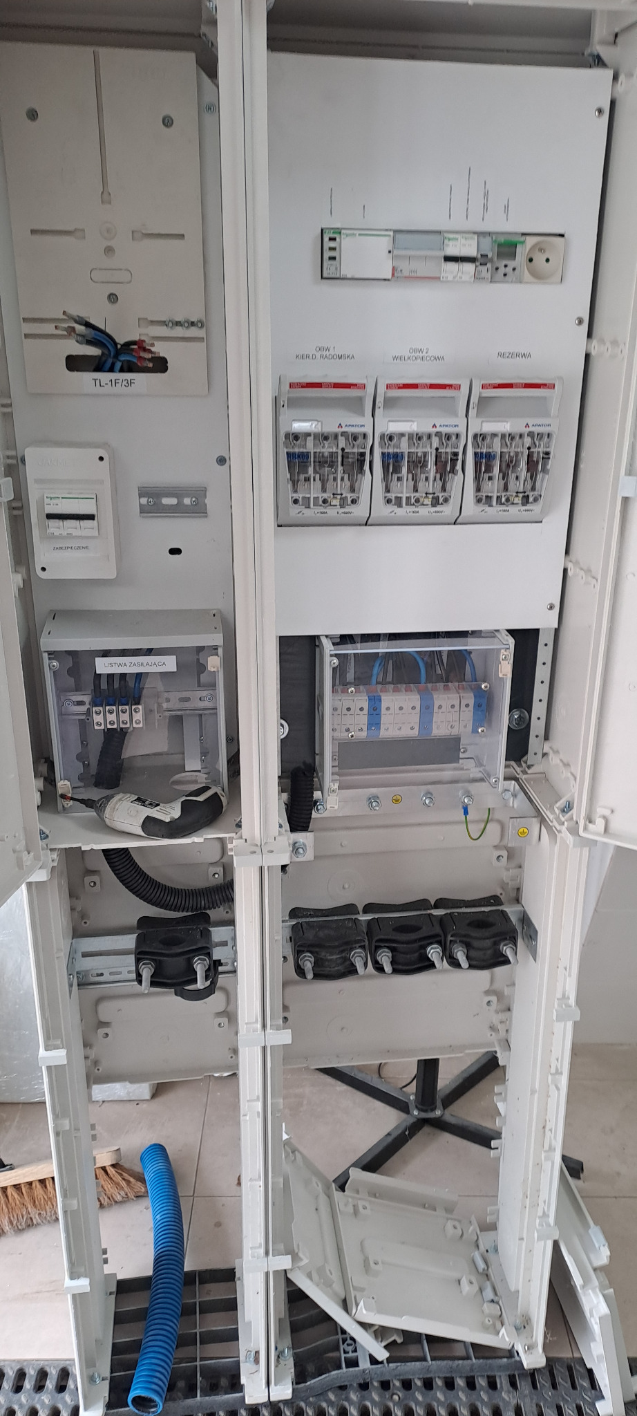 Otwarta szafa elektryczna w trakcie montażu, widoczne zabezpieczenia, listwy zasilające, przewody i narzędzia, w tle podłoga i rura karbowana.
