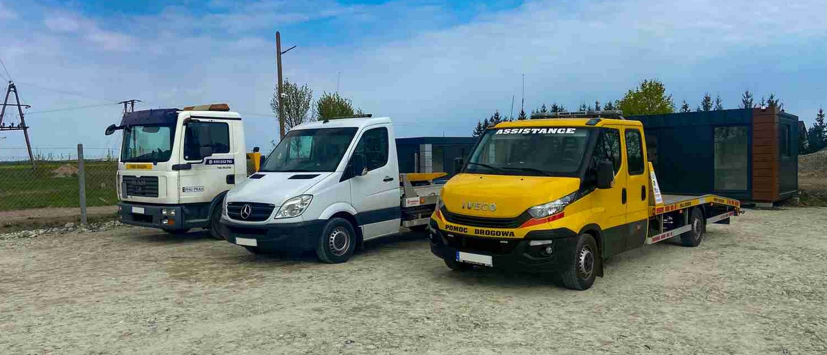 Trzy pojazdy pomocy drogowej: biały Mercedes Sprinter, żółty Iveco z napisem 'Assistance' oraz biały MAN, stoją zaparkowane na żwirowym placu.