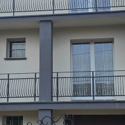 VITALII ORDASH-BUD - Dwupiętrowy budynek z balkonami o szarej balustradzie i kolumnach. Elewacja w jasnym odcieniu, okna z białymi ramami. Widoczne fragmenty dachu i drzewa w tle.