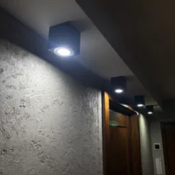 Rząd czterech kwadratowych, wpuszczanych lamp sufitowych LED oświetlających korytarz z teksturowaną ścianą i drewnianymi drzwiami.