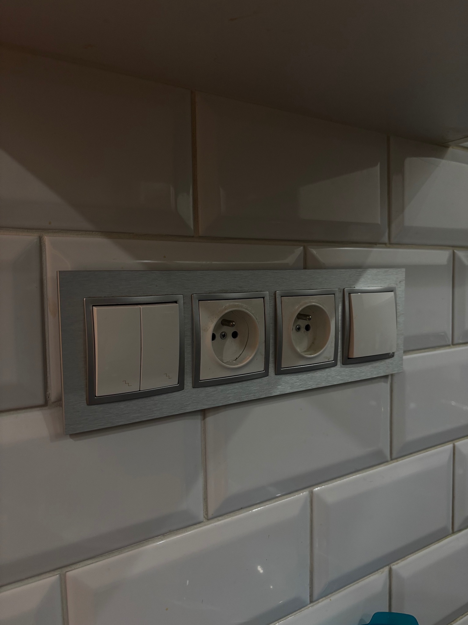 Panel z dwoma włącznikami światła i dwoma gniazdkami elektrycznymi, zamontowany na ścianie wyłożonej białymi, podłużnymi płytkami.