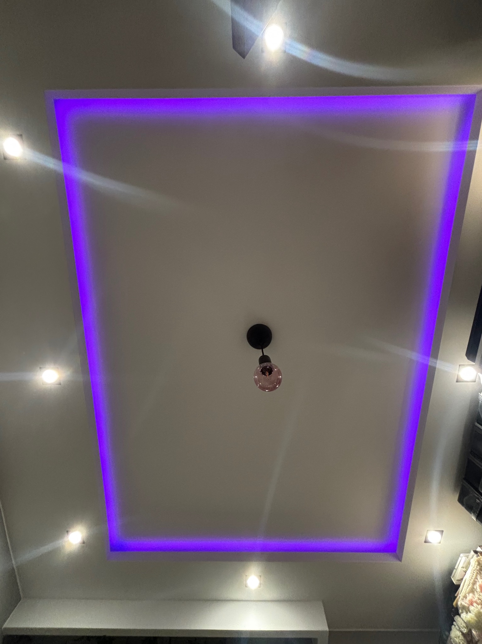 Nowoczesny sufit podwieszany z oświetleniem LED w kolorze fioletowym, centralnie umieszczona lampa wisząca z różową żarówką i dodatkowe punktowe oświetlenie.