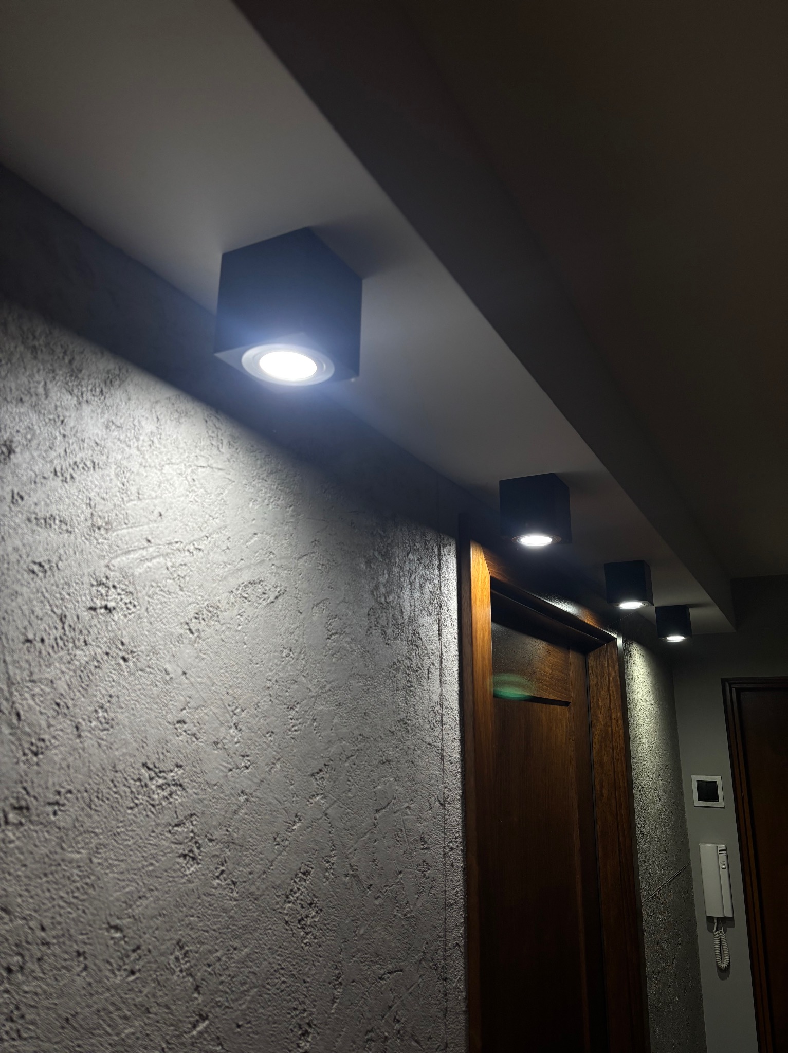 Rząd czterech kwadratowych, wpuszczanych lamp sufitowych LED oświetlających korytarz z teksturowaną ścianą i drewnianymi drzwiami.