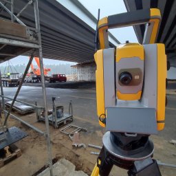 ALEGEO Usługi Geodezyjne Aleksandra Józefowicz-Stasiak - Żółto-szary tachimetr Trimble na statywie, na tle budowy mostu z dźwigiem i rusztowaniami, widok z bliska, lekko pochmurna pogoda.