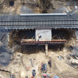 ALEGEO Usługi Geodezyjne Aleksandra Józefowicz-Stasiak - Zdjęcie z drona przedstawia budowę betonowego przepustu pod linią kolejową. Widoczny pracownik w pomarańczowym kasku na betonowej płycie, drewniane szalunki, wykop i linia kolejowa z podkładami.