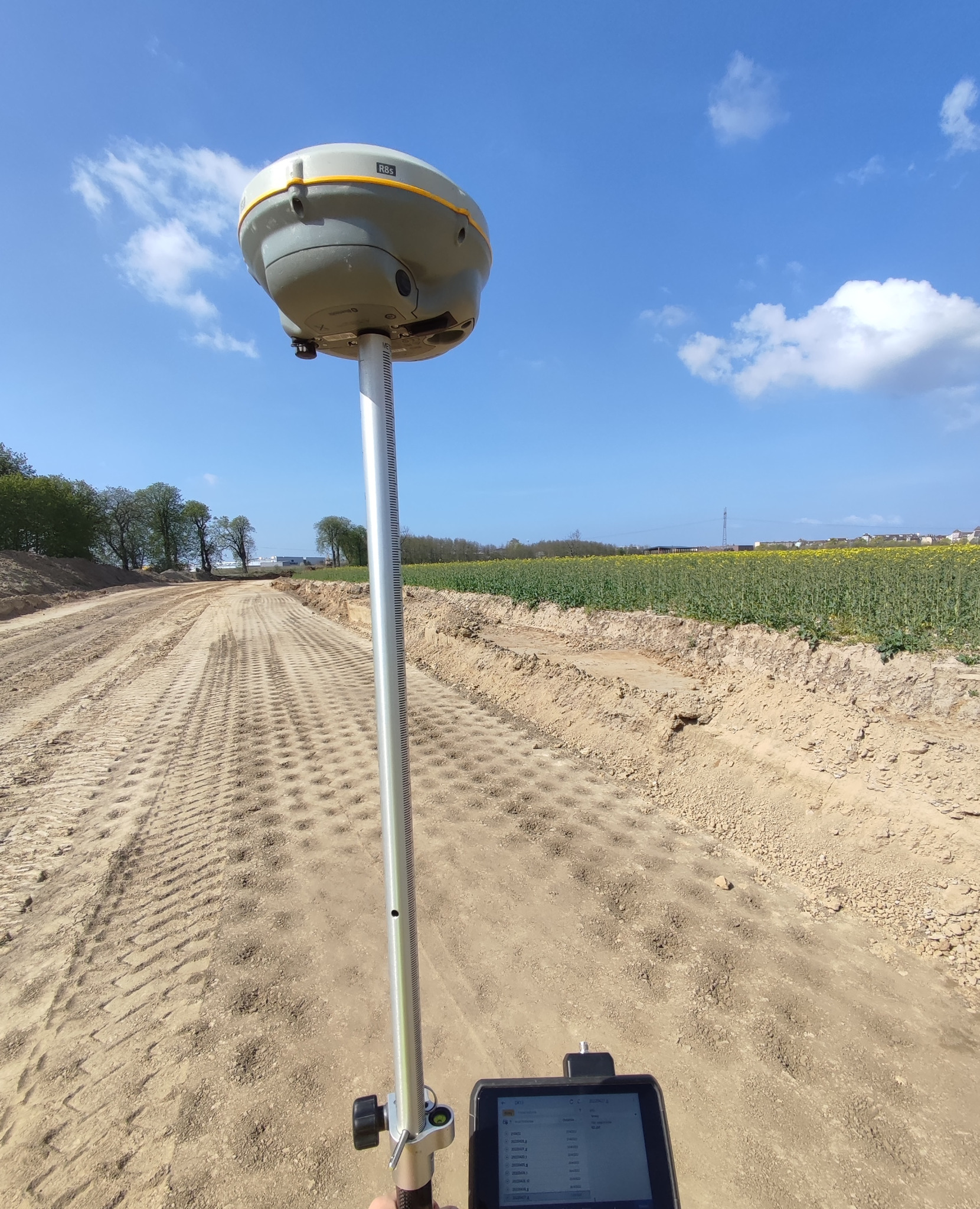 Pomiar terenu budowy drogi za pomocą odbiornika GNSS RTK na tyczce, z widocznym tabletem sterującym i polami w tle pod błękitnym niebem.