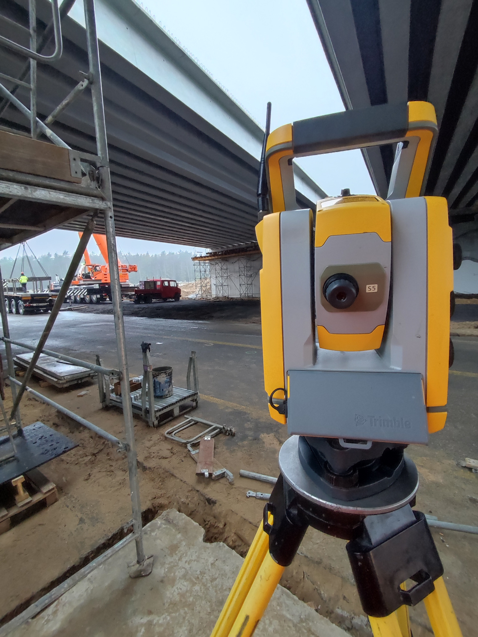Żółto-szary tachimetr Trimble na statywie, na tle budowy mostu z dźwigiem i rusztowaniami, widok z bliska, lekko pochmurna pogoda.