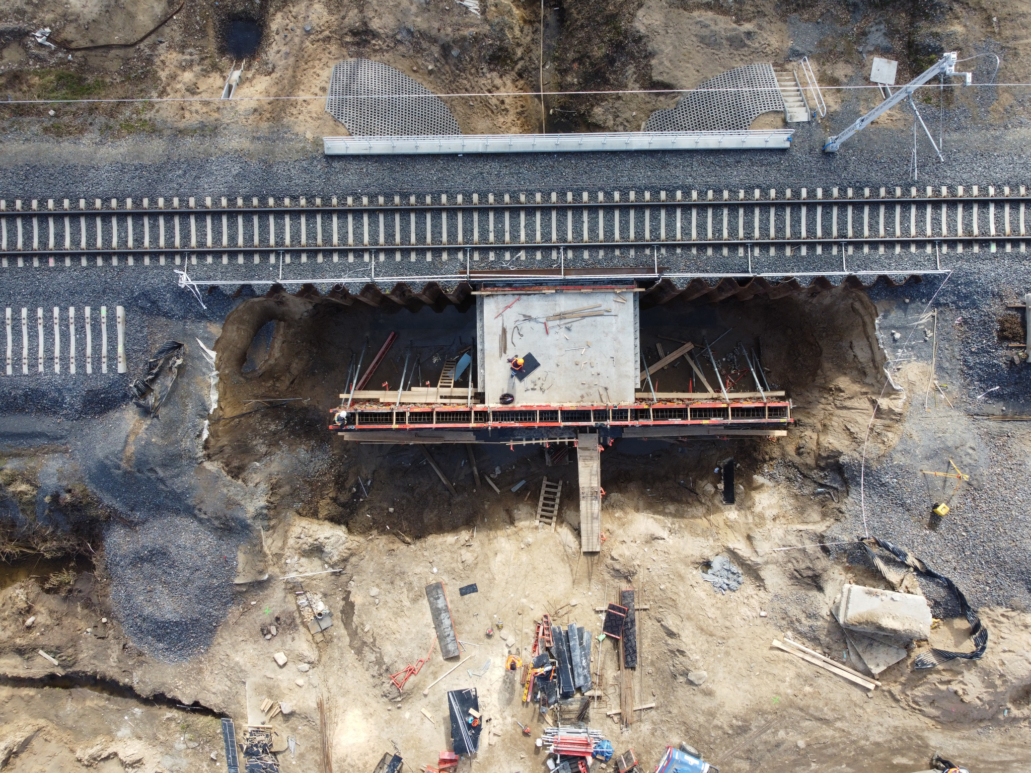 Zdjęcie z drona przedstawia budowę betonowego przepustu pod linią kolejową. Widoczny pracownik w pomarańczowym kasku na betonowej płycie, drewniane szalunki, wykop i linia kolejowa z podkładami.