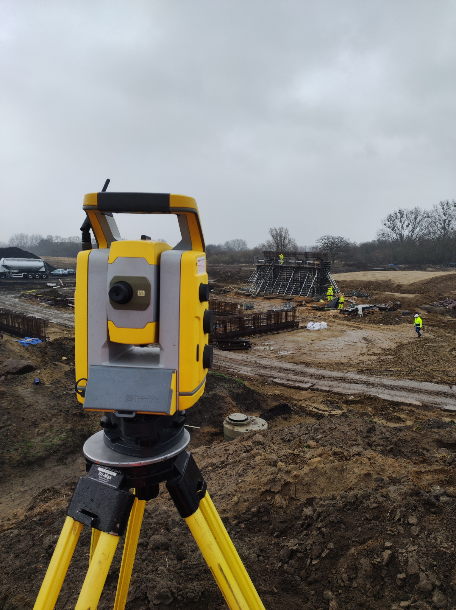 Żółty tachimetr Trimble na statywie na placu budowy z widocznymi pracownikami w odblaskowych kamizelkach i konstrukcją zbrojeniową w tle, pochmurny dzień.