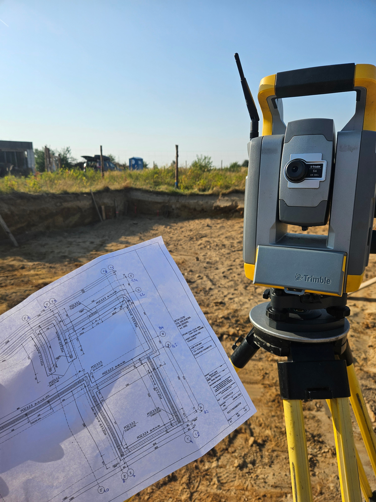 Szkic fundamentów trzymany na tle żółto-szarego teodolitu Trimble na trójnogu, w tle wykop pod fundamenty i błękitne niebo.