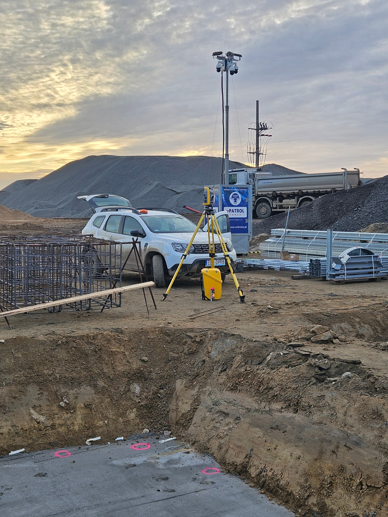 Żółty niwelator optyczny ustawiony na statywie obok białego samochodu Dacia Duster na placu budowy z widocznymi zbrojeniami, wykopem i hałdą piasku w tle.