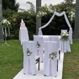 Plenerowa aranżacja ślubna: biały dywan, krzesła z białymi pokrowcami i kwiatowymi dekoracjami, złoty łuk z białymi draperiami i kwiatami, kolumny w tle, na trawniku, z widocznym godłem Polski w tle.