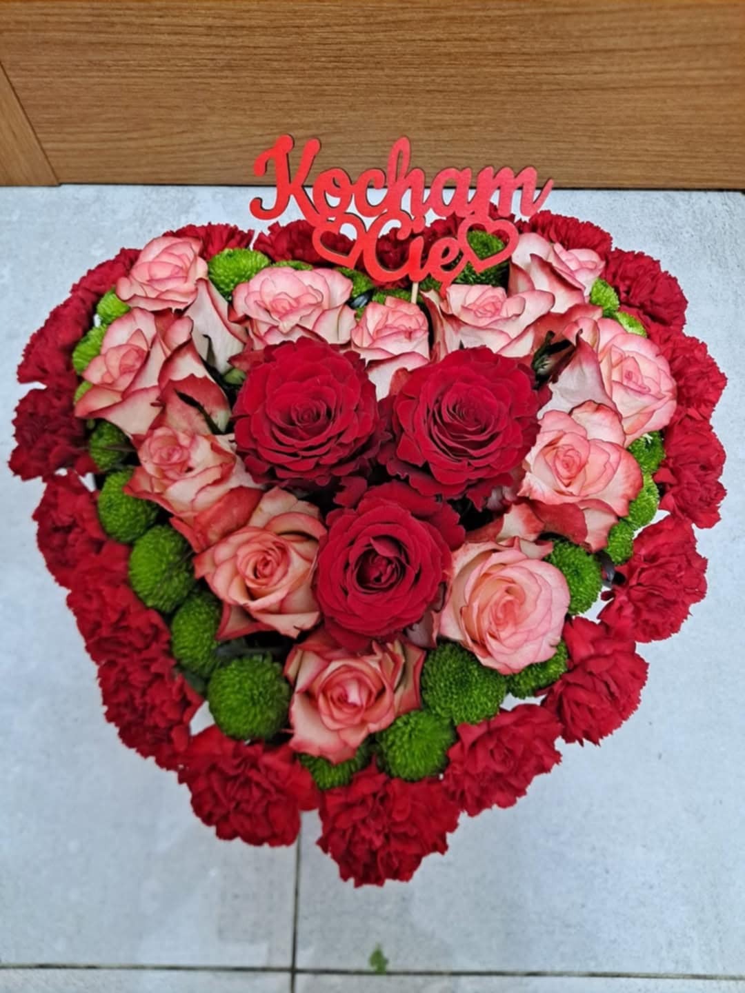 Kompozycja kwiatowa w kształcie serca z czerwonych i różowych róż, czerwonych goździków oraz zielonych chryzantem, z czerwonym napisem 'Kocham Cię' na górze.