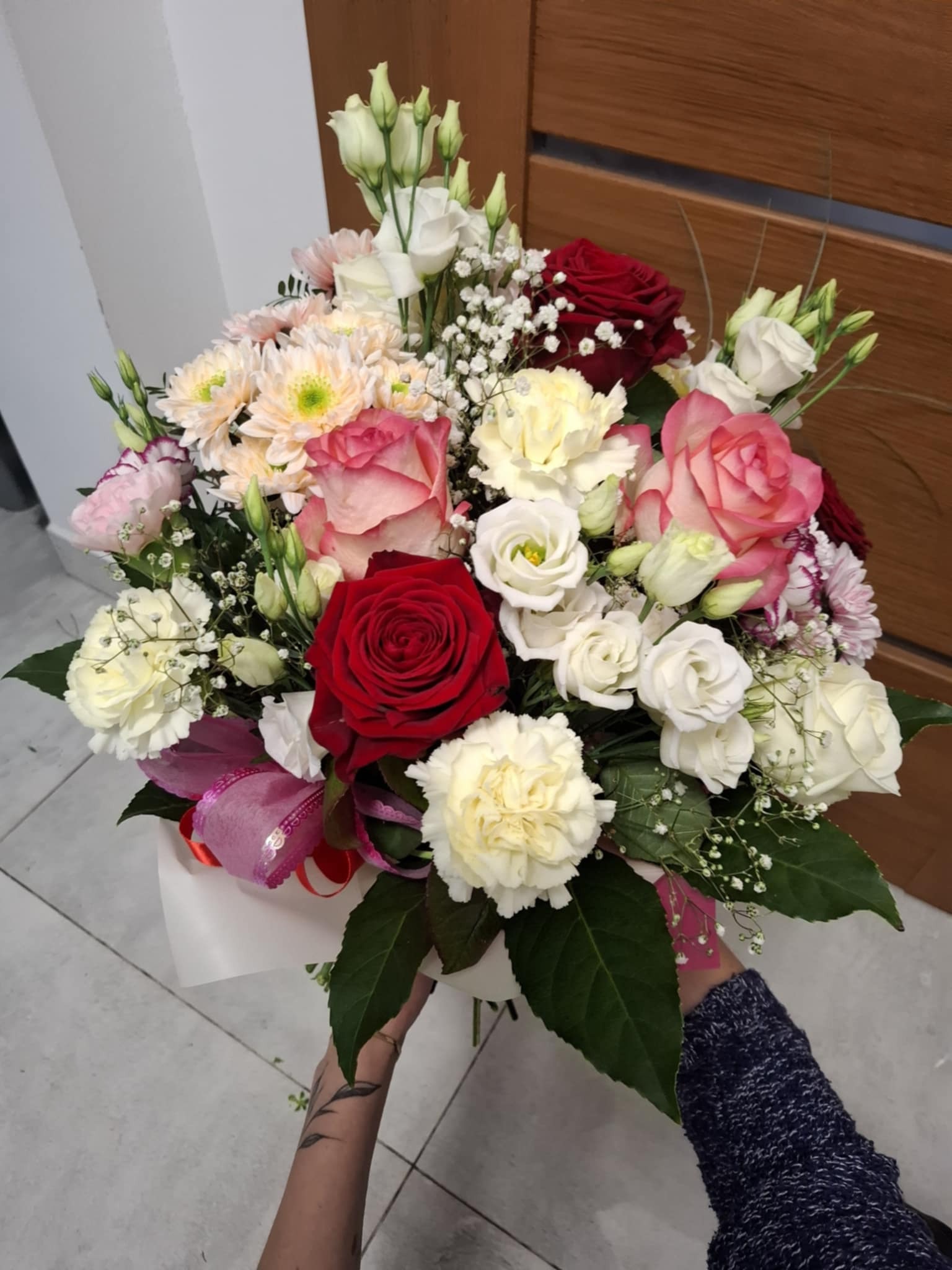 Bukiet mieszanych kwiatów w odcieniach czerwieni, różu i bieli, trzymany w dłoniach osoby z tatuażem na przedramieniu, na tle jasnej ściany i drewnianych drzwi.