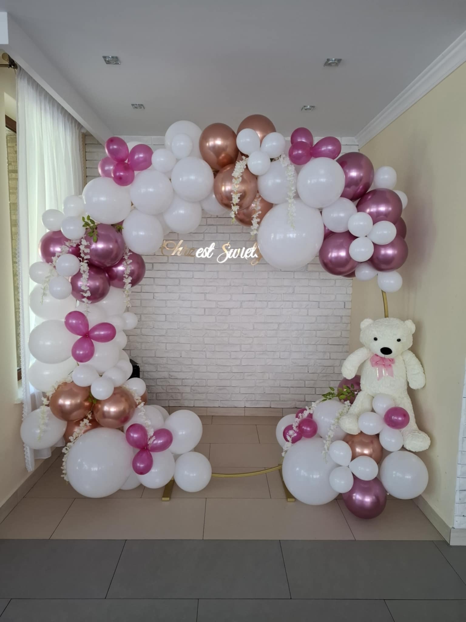 Dekoracja balonowa z misiem w Krasnosielcu: białe, różowe i złote balony tworzą łuk przed białą ceglaną ścianą, ozdobiony drobnymi kwiatkami.