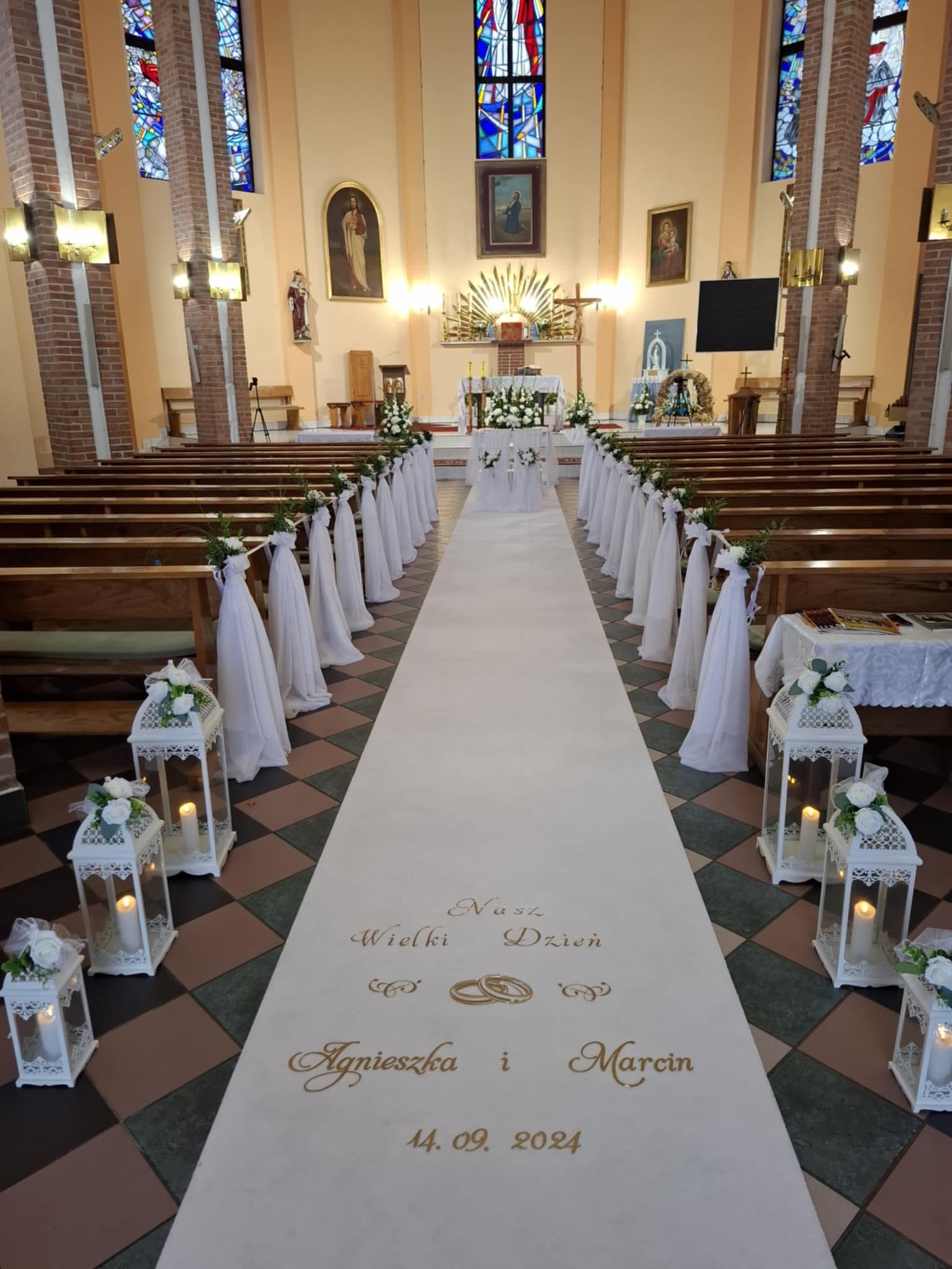 Uroczystość ślubna w kościele: biały dywan z imionami Agnieszka i Marcin oraz datą 14.09.2024, ustawiony wzdłuż nawy głównej, otoczony drewnianymi ławkami udekorowanymi białymi tkaninami i kwiatami...