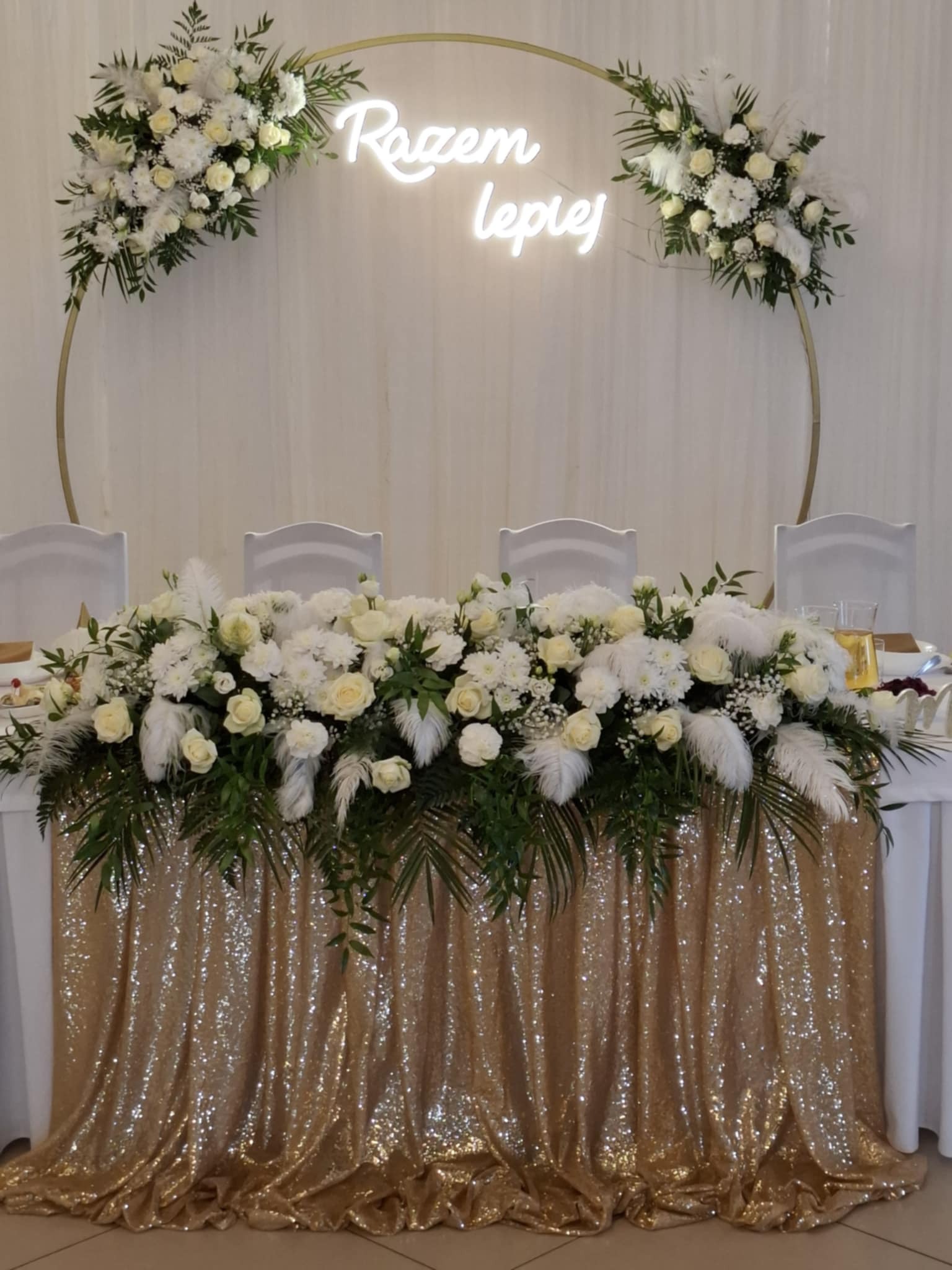 Dekoracja stołu weselnego z białych kwiatów, piór i zielonych liści, z błyszczącą złotą tkaniną i neonowym napisem 'Razem Lepiej' w tle.