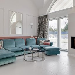 Madro Real Estate Szczecin - Kreatywna sprzedaż nieruchomości - Pas nadmorski - Jasne, przestronne wnętrze salonu z turkusową sofą, okrągłymi stolikami kawowymi, kominkiem z palącym się ogniem i dużym oknem z widokiem na ośnieżony ogród.